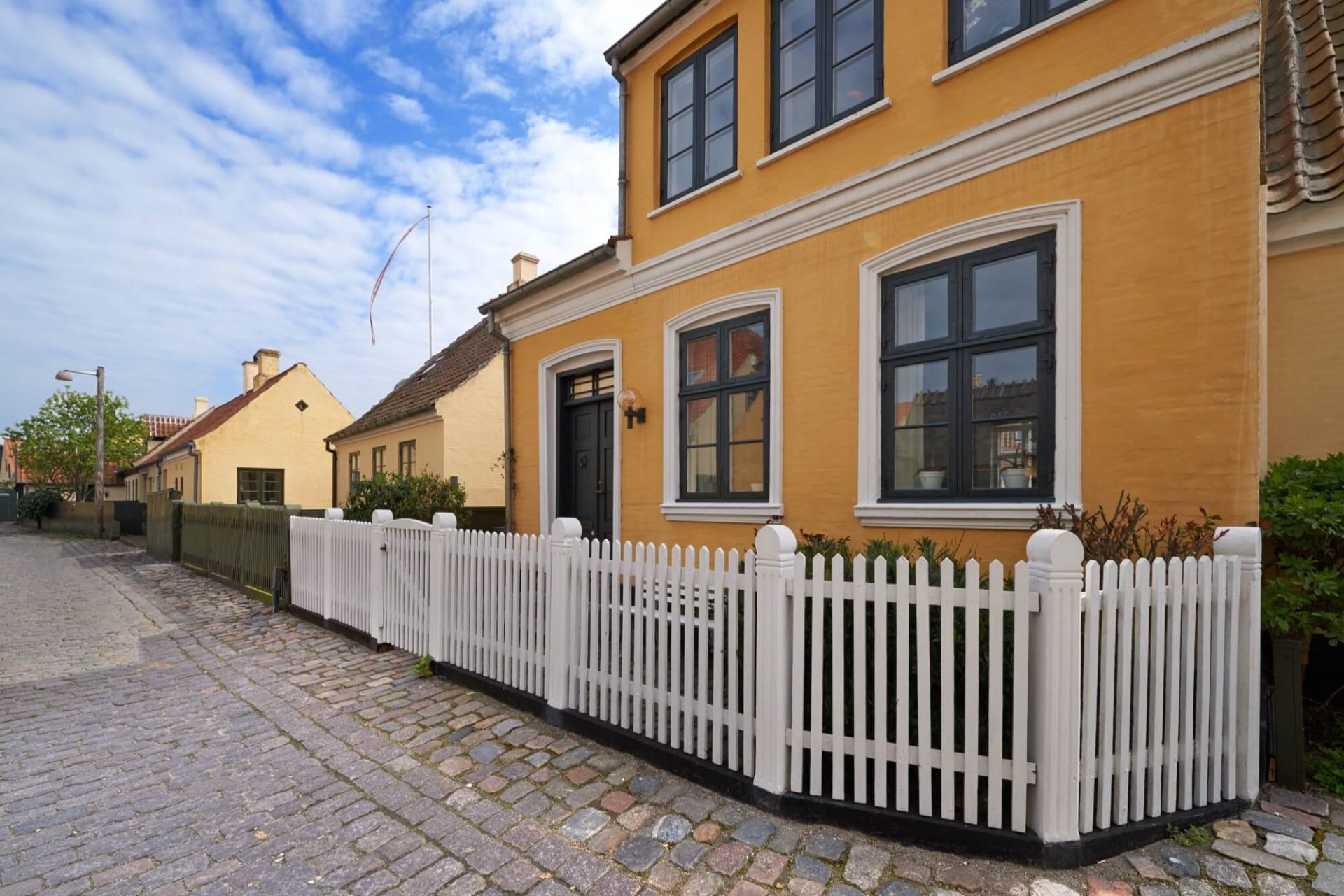 Ett traditionellt hus med en markant gul yttre, fönster iklädda svart och ett staket i vitt trä, framträder längs en gata som är vackert belagd med kullersten