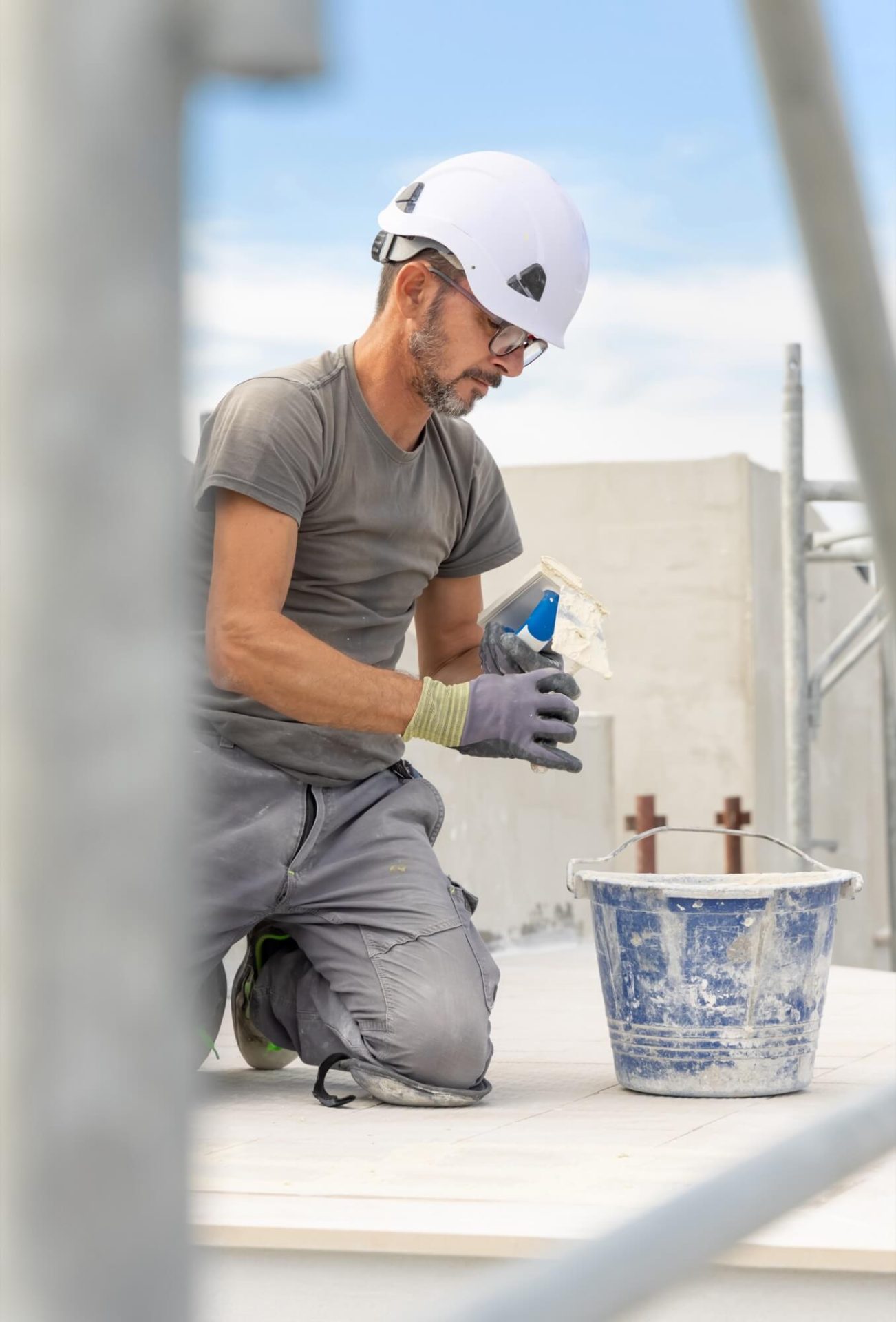 Medan han är knästående på marken, utför en hantverkare med skyddshjälm och handskar en noggrann fasadrenovering, omgiven av sitt murbruk och verktyg